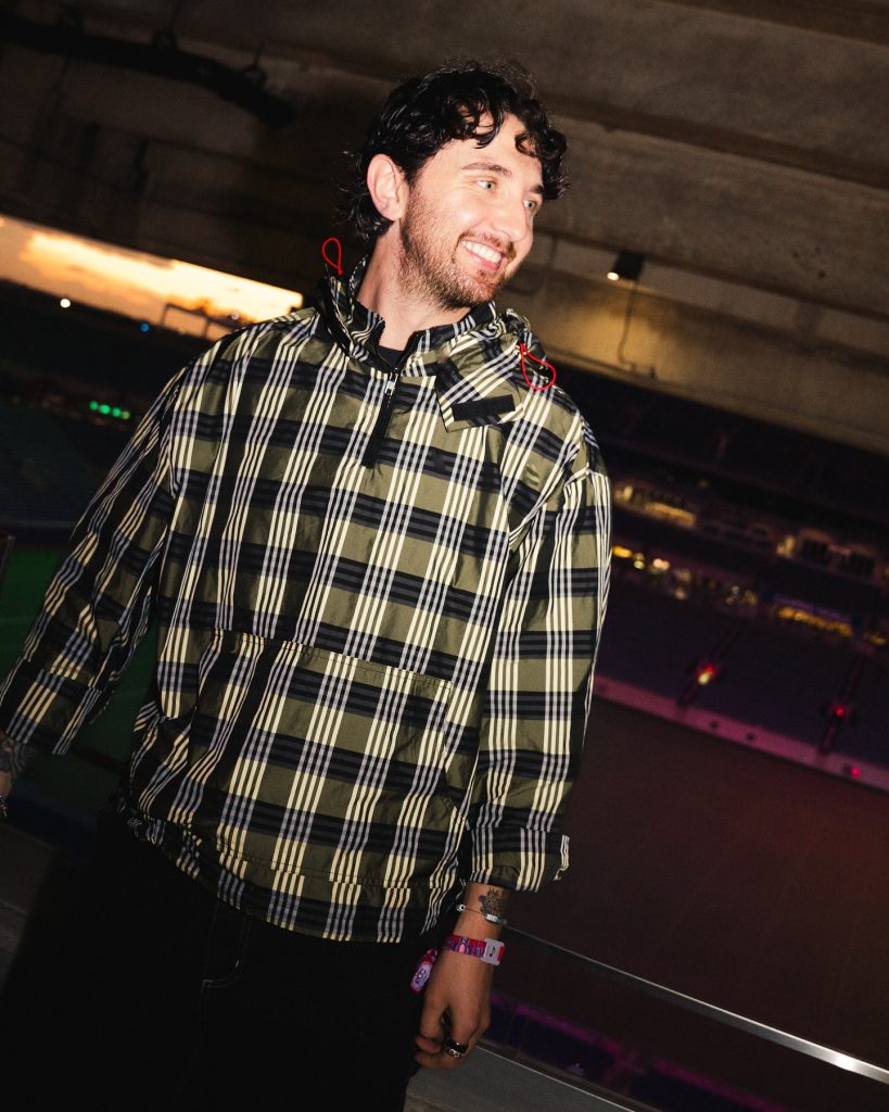 "Retrato de Max Styler sonriendo y mirando hacia un lado en un entorno nocturno, vistiendo una chaqueta impermeable con patrón de cuadros en tonos verdes y negros."