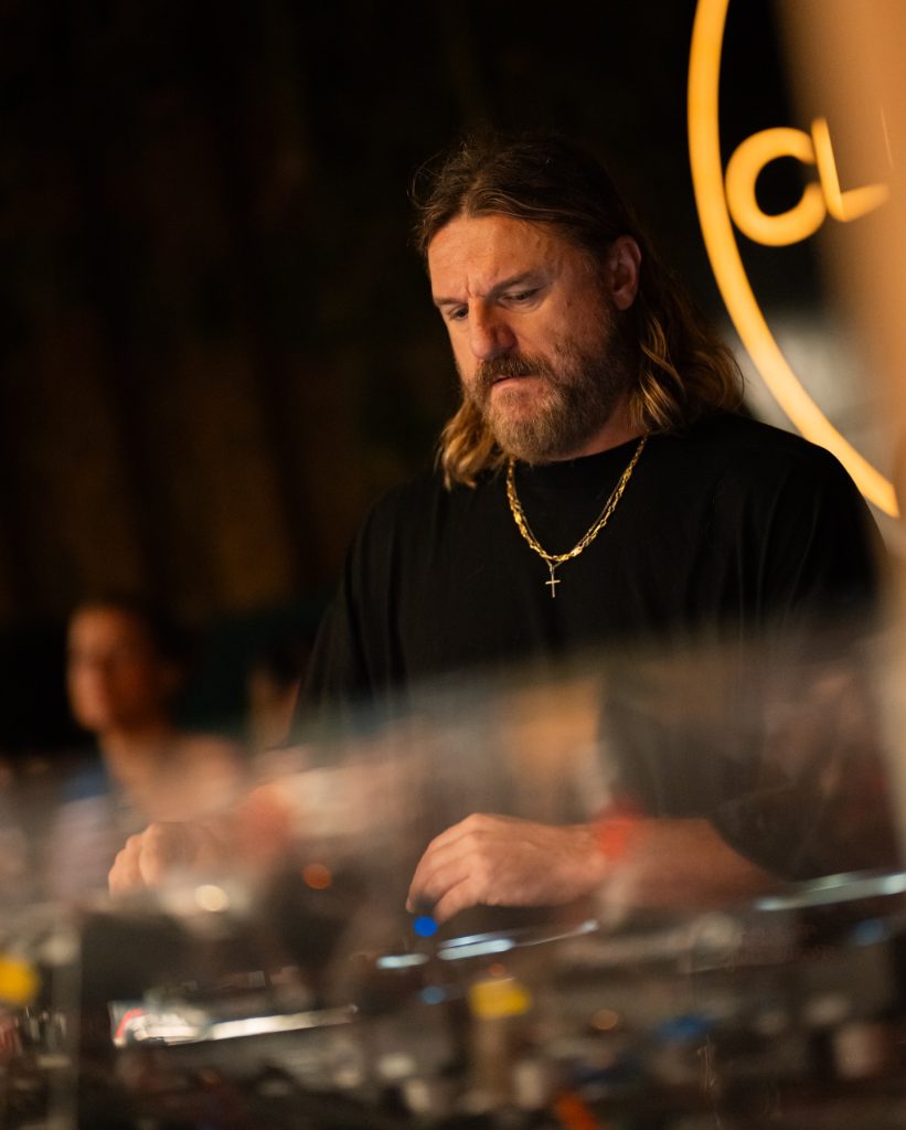 "Primer plano del DJ Solomun mezclando en vivo bajo iluminación cálida, vistiendo camiseta negra y cadena dorada, enfocado en la consola durante un set nocturno."