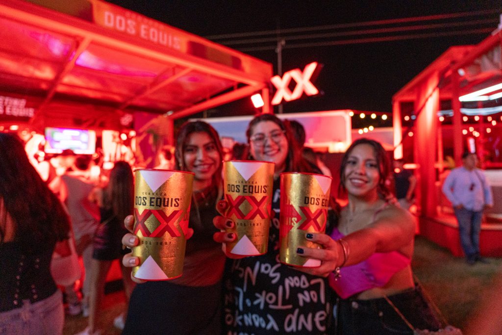 Grupo de tres amigas sonriendo y haciendo un brindis hacia la cámara con sus vasos de cerveza Dos Equis en un ambiente de fiesta con luces rojas.