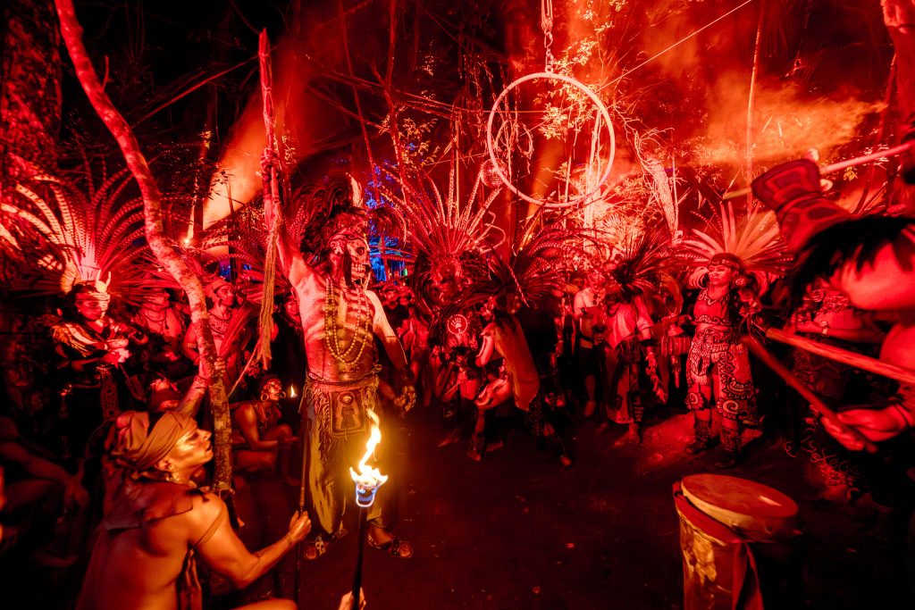eremonia nocturna en la selva durante el festival Day Zero, con artistas vestidos con elaborados atuendos y penachos mayas, pintura corporal y antorchas, bajo una intensa iluminación roja.