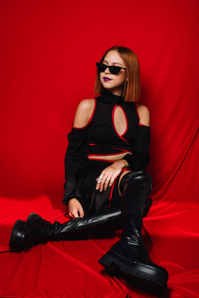 Retrato de DANGR en estudio, de perfil, usando lentes oscuros y top negro sin mangas, con fondo neutro claro que resalta su estilo sobrio y minimalista.