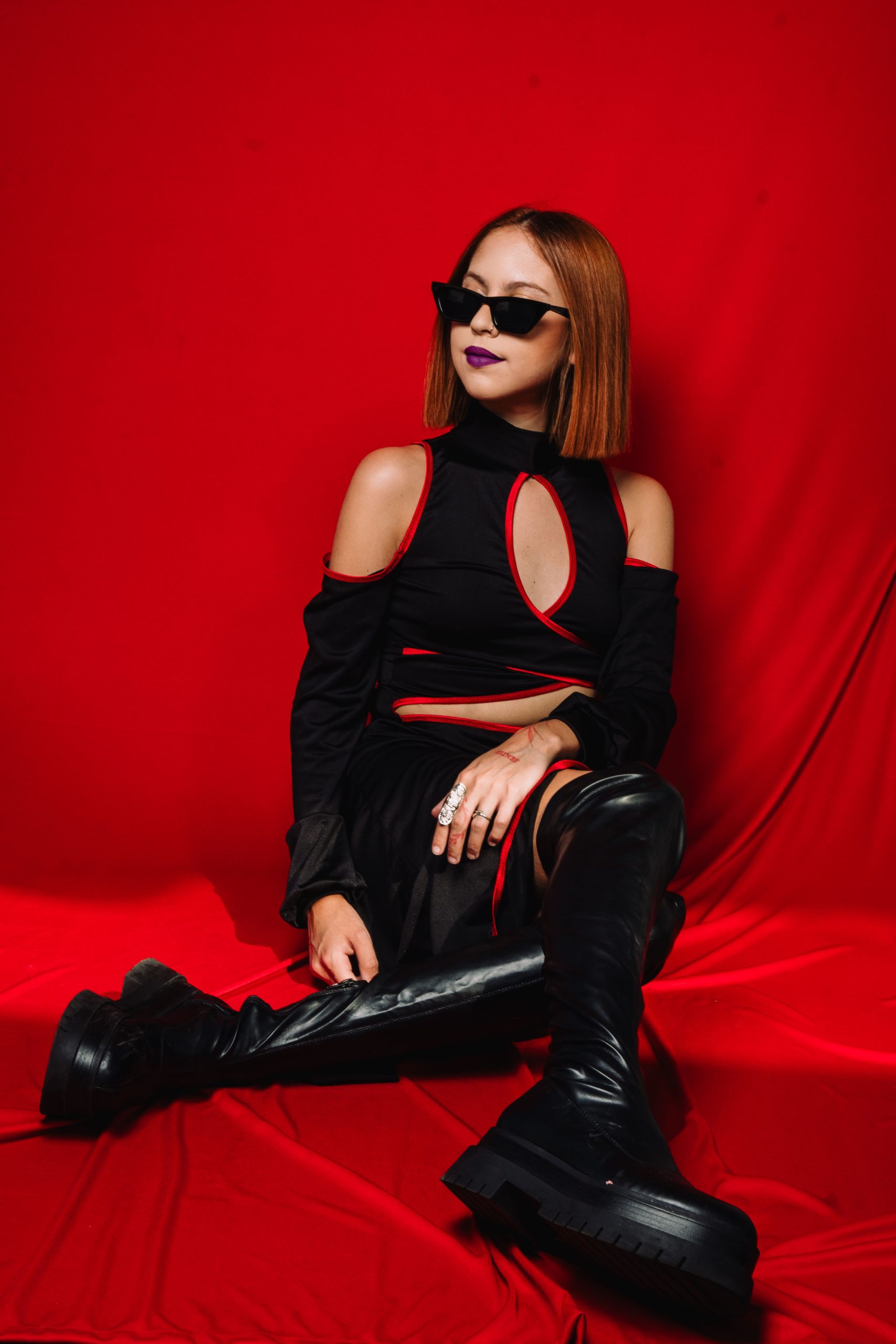 Retrato de DANGR en estudio, de perfil, usando lentes oscuros y top negro sin mangas, con fondo neutro claro que resalta su estilo sobrio y minimalista.