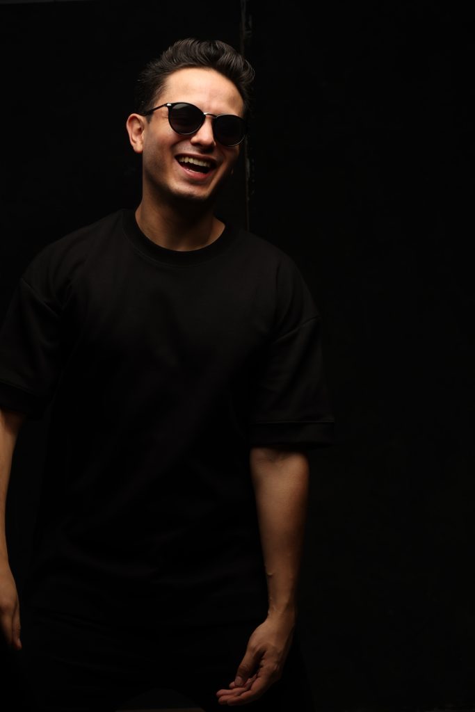 BZARS sonríe frente a un fondo oscuro, vestido completamente de negro, en una sesión fotográfica de estudio.