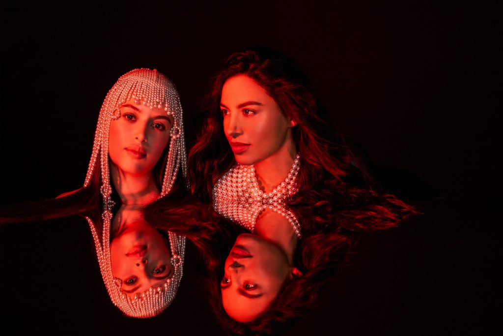 D’Witches en retrato doble con estilismo oscuro y accesorios metálicos, reflejadas en una superficie pulida con iluminación dramática roja.