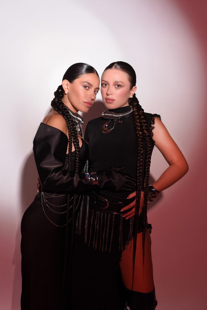 Las hermanas Thania y Thamara Ortiz posando juntas con vestuario negro y peinados trenzados, en una sesión editorial de estudio.
