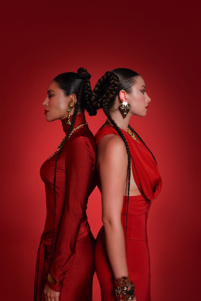 D’Witches de perfil, espalda con espalda, vestidas en tonos rojos sobre fondo monocromático, reforzando la estética ritual del dúo.