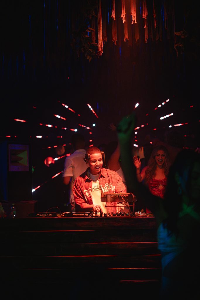 Dezko mezclando en club nocturno con iluminación roja y azul, rodeado de público en ambiente íntimo.