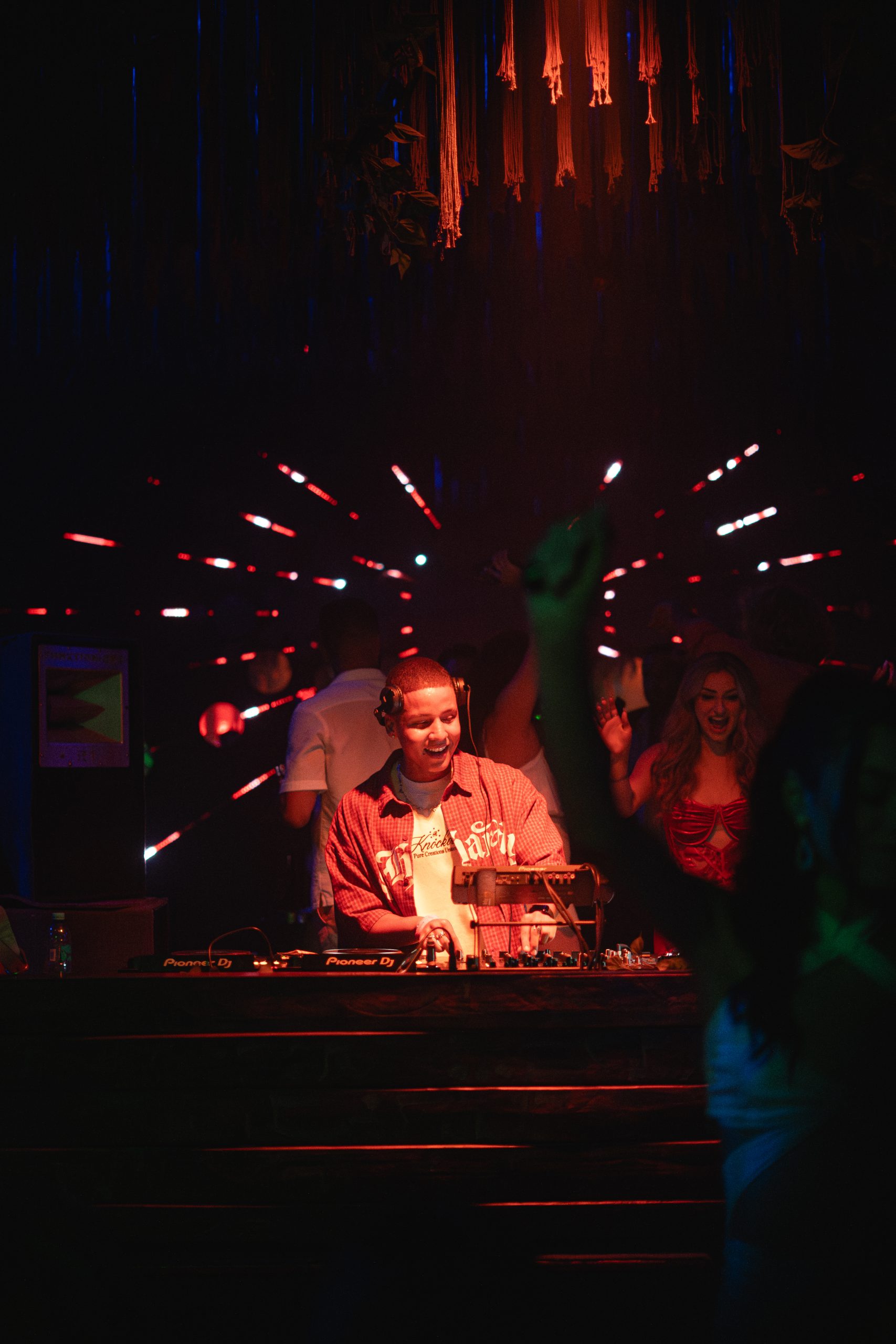 Dezko mezclando en club nocturno con iluminación roja y azul, rodeado de público en ambiente íntimo.