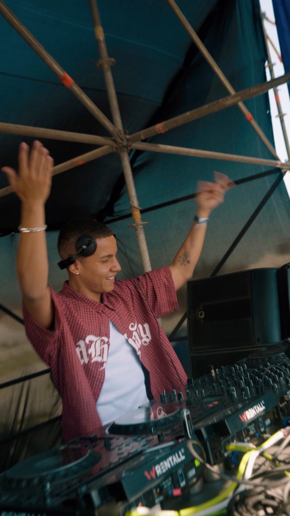 Dezko levantando las manos durante un DJ set al aire libre, frente a cabina profesional y estructura técnica de escenario.