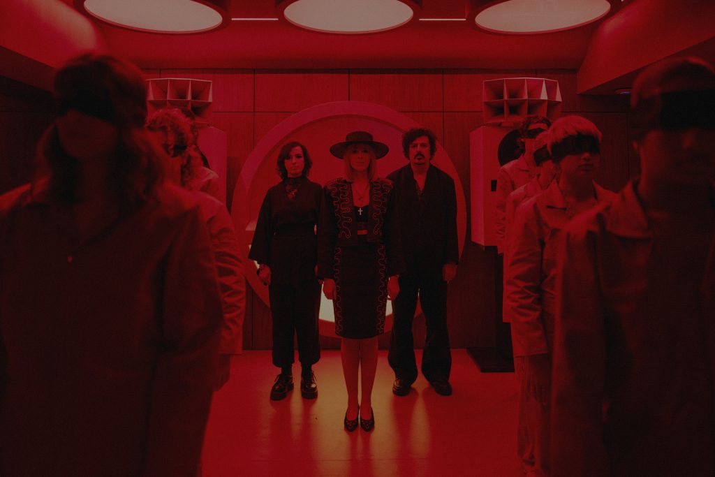 Ladytron de pie en una sala iluminada en rojo con figuras humanas desenfocadas, imagen conceptual que acompaña la narrativa visual del single “A Death In London”.