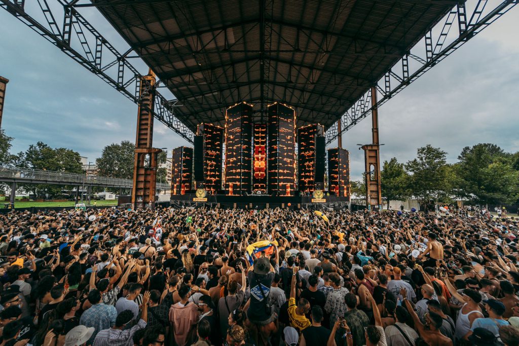 escenario principal bajo techo industrial
Escenario principal de Kappa Futur Festival visto desde el fondo de una multitud con los brazos en alto. La estructura metálica industrial sirve de techo al espacio. El escenario muestra una pantalla LED de gran formato con iluminación cálida en tonos dorado y naranja.