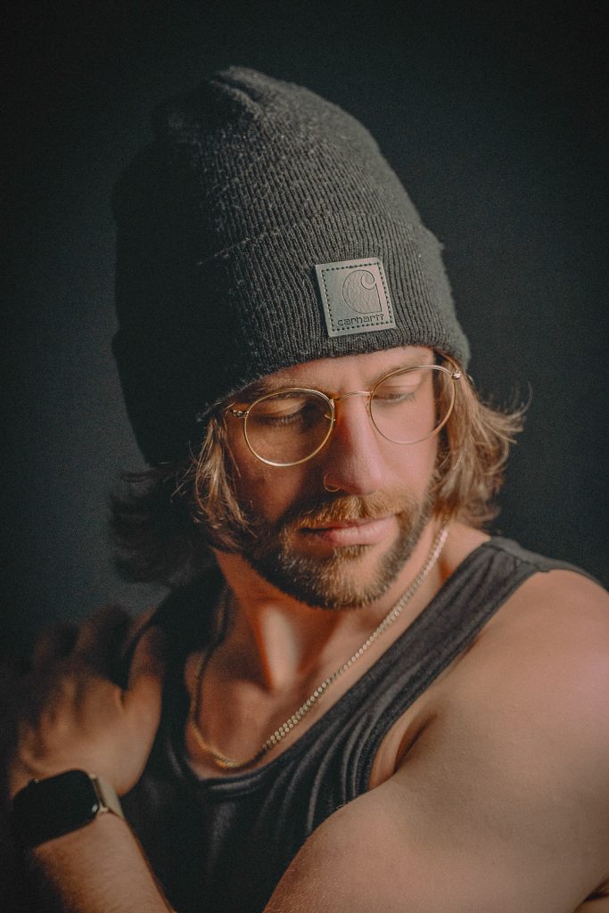 ivingston Crain, vocalista de EDM y pop-punk, con lentes aviador y gorro gris, sobre fondo oscuro.