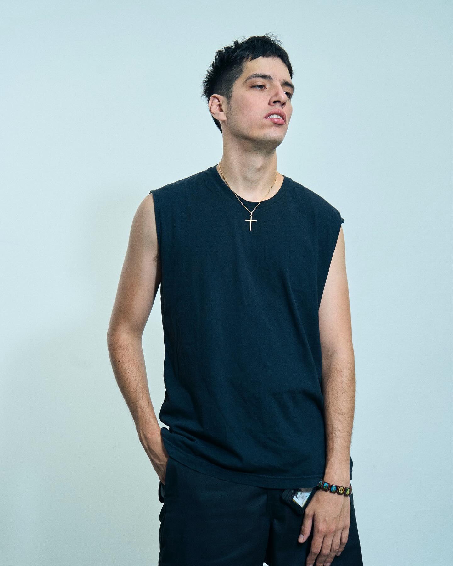 Retrato de estudio del DJ y productor Mandragora, con playera negra sin mangas, collar con cruz y pulsera de colores, mirando hacia arriba con actitud relajada.