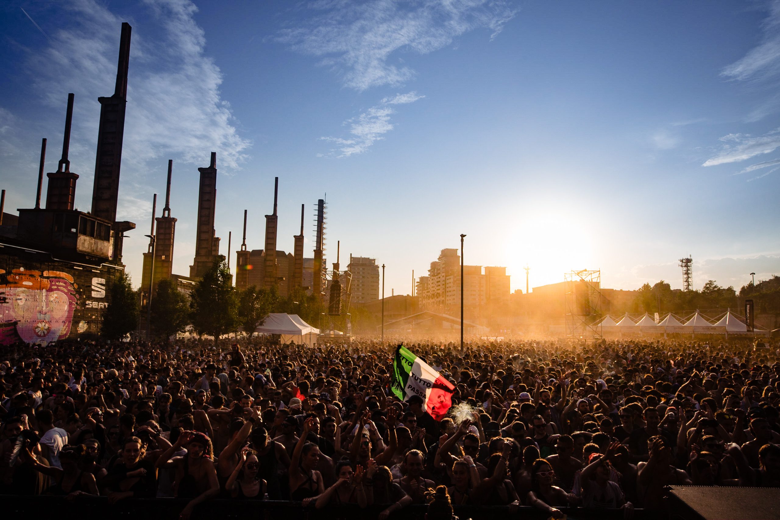 multitud en Kappa Futur Festival, Turín al atardecer Mar de personas en el Parco Dora de Turín durante el atardecer en una edición de Kappa Futur Festival. Las chimeneas industriales se recortan en silueta contra el sol poniente y el cielo despejado. Alguien en el centro de la multitud agita banderas de colores verde, rojo y naranja.