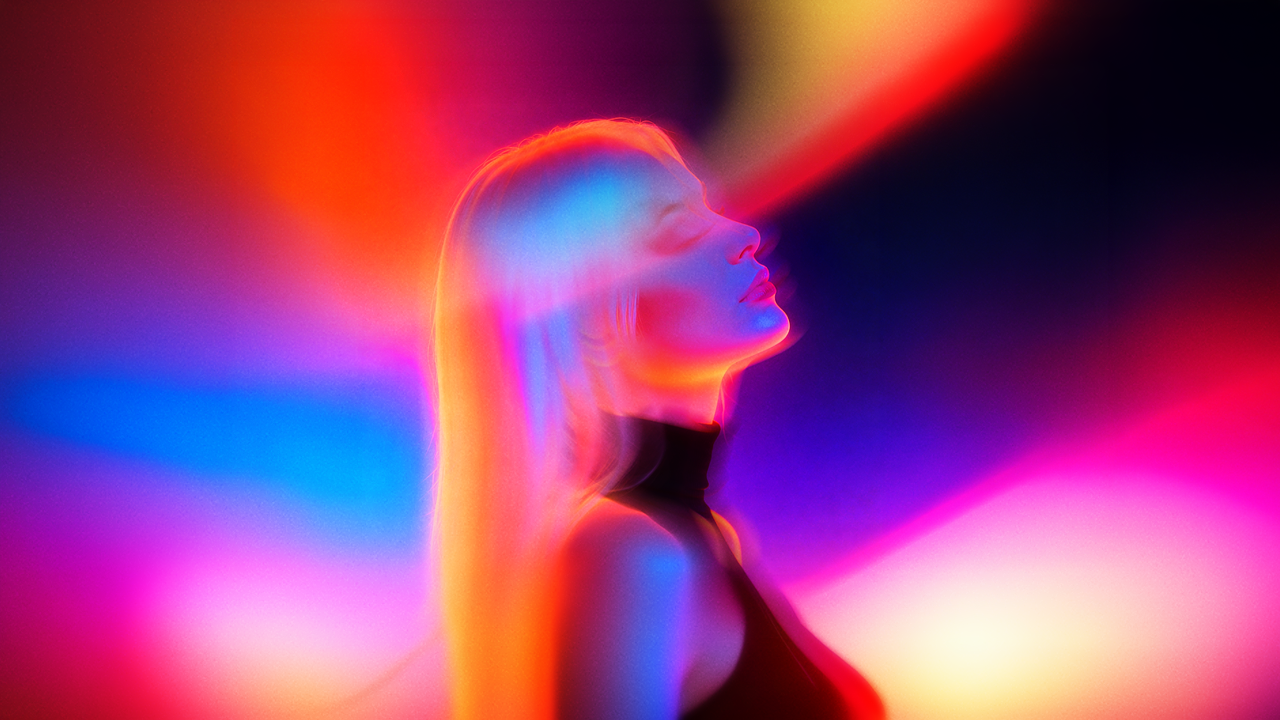 Fotografía artística de perfil de mujer con cabello largo rubio, cabeza inclinada hacia atrás con ojos cerrados. Imagen intencionalmente difuminada con luces de colores vibrantes — azul, naranja, rosa y rojo — que envuelven la silueta. Fondo abstracto con rayos de luz neón sobre negro.
