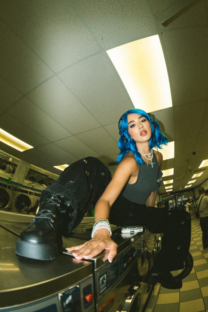 CiCiBi con cabello azul en sesión fotográfica editorial dentro de una lavandería, estética alternativa y urbana.