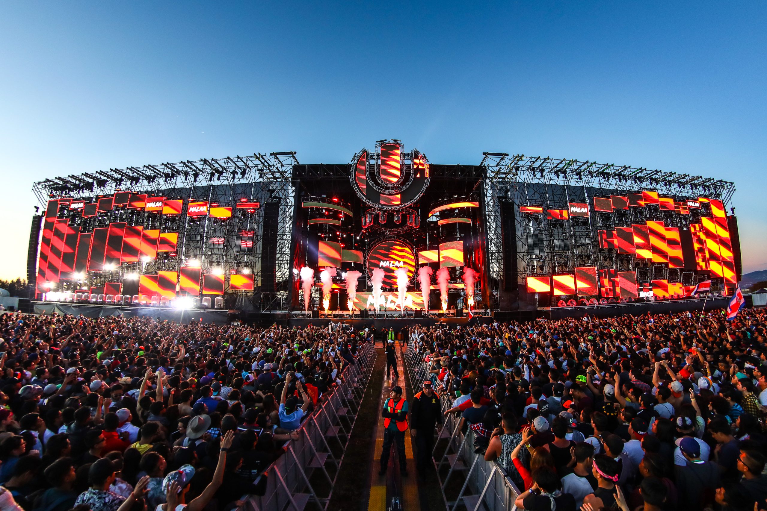 Vista panorámica del escenario principal de Ultra México durante el día, mostrando una imponente estructura repleta de pantallas LED con gráficos en rojo y amarillo, columnas de humo blanco y miles de fans frente al escenario con las manos levantadas.