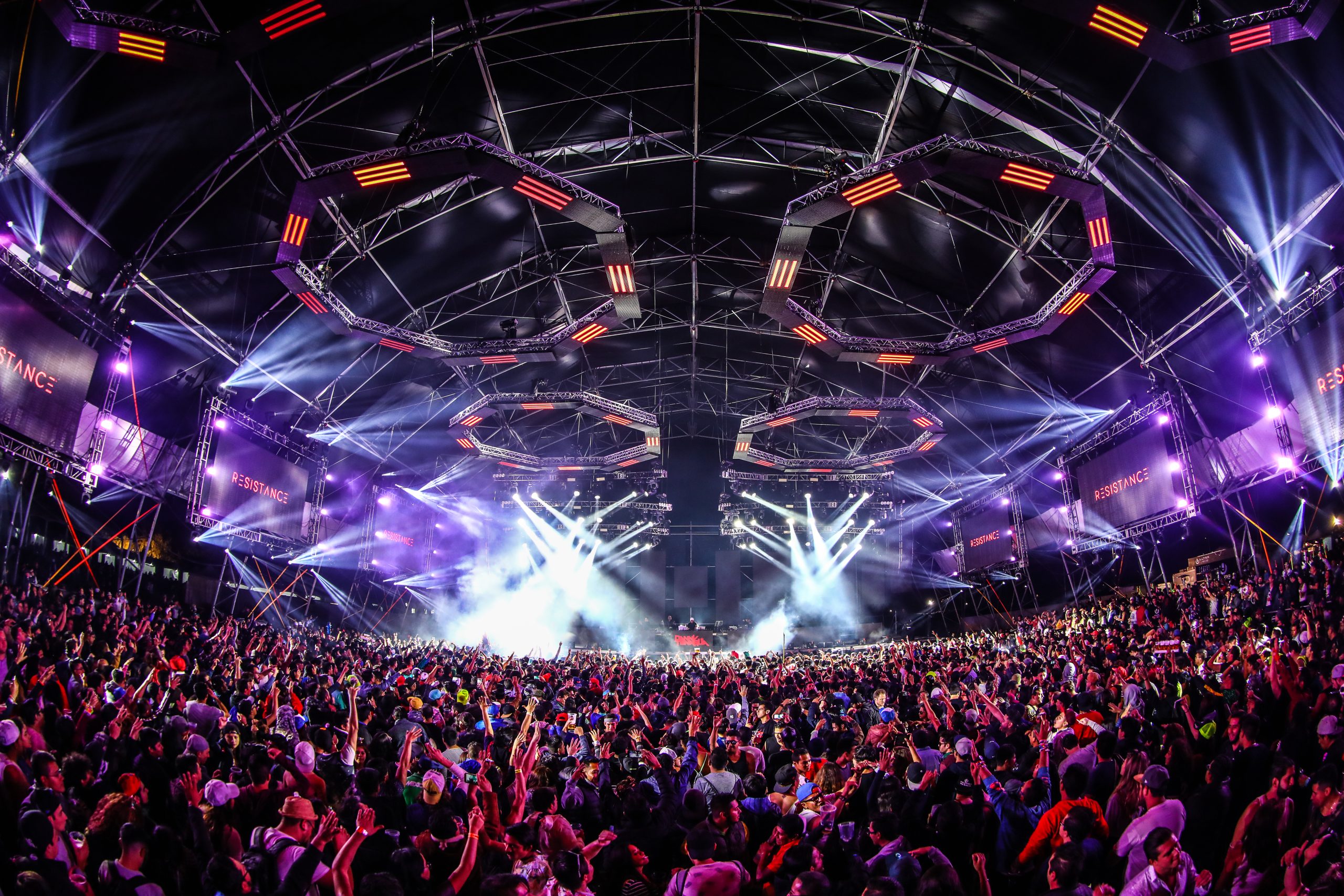 Fotografía nocturna del escenario Resistance bajo una carpa industrial masiva, con estructuras hexagonales iluminadas en rojo en el techo, pantallas gigantes laterales, luces de haz blanco y miles de asistentes disfrutando de la música techno y house.