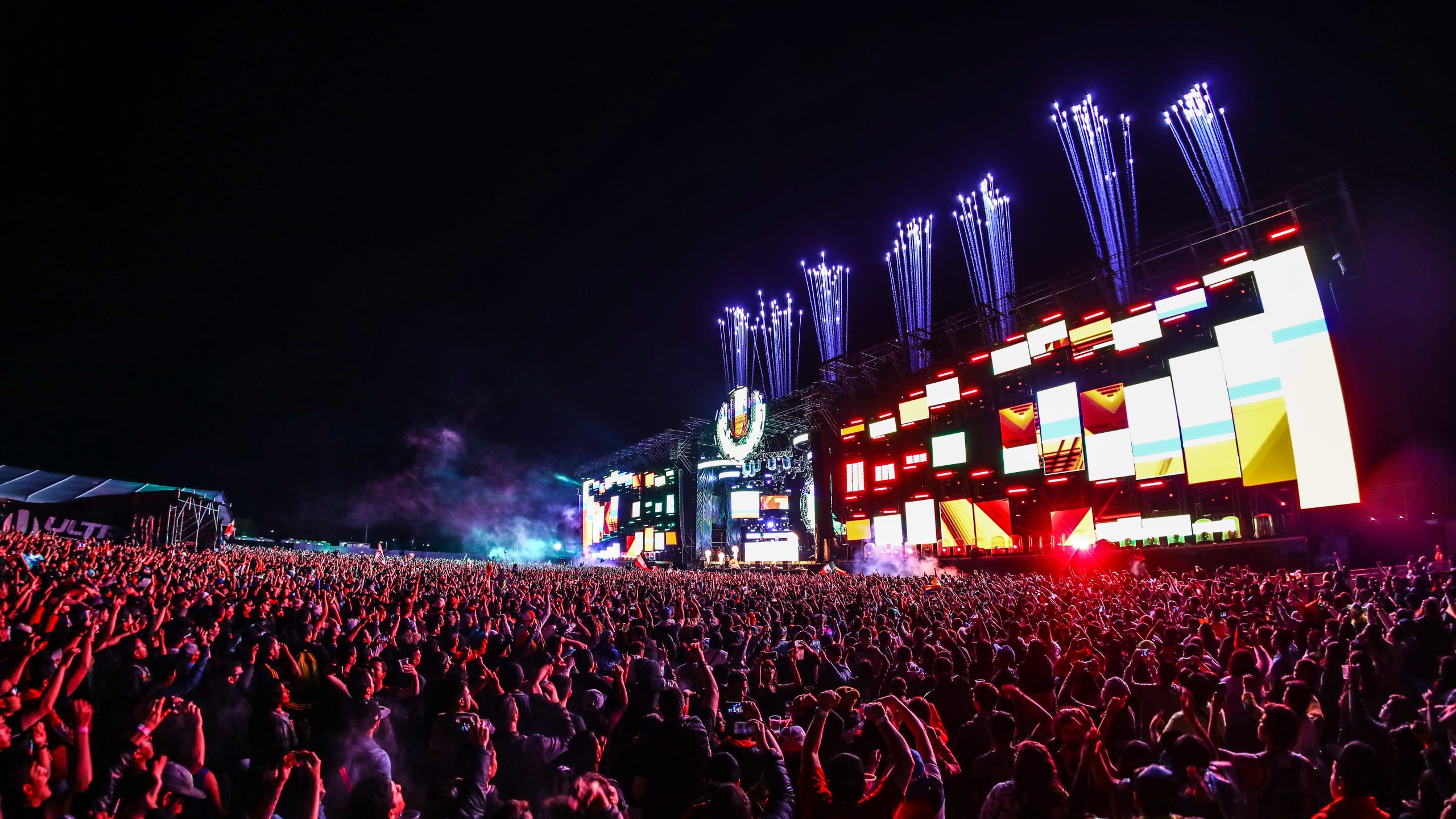 Tomas nocturnas del Ultra Main Stage en pleno show, con pantallas LED encendidas en tonos rojos, una multitud masiva iluminada por la luz del escenario y fuegos artificiales de chispas blancas disparados desde la cima del escenario.