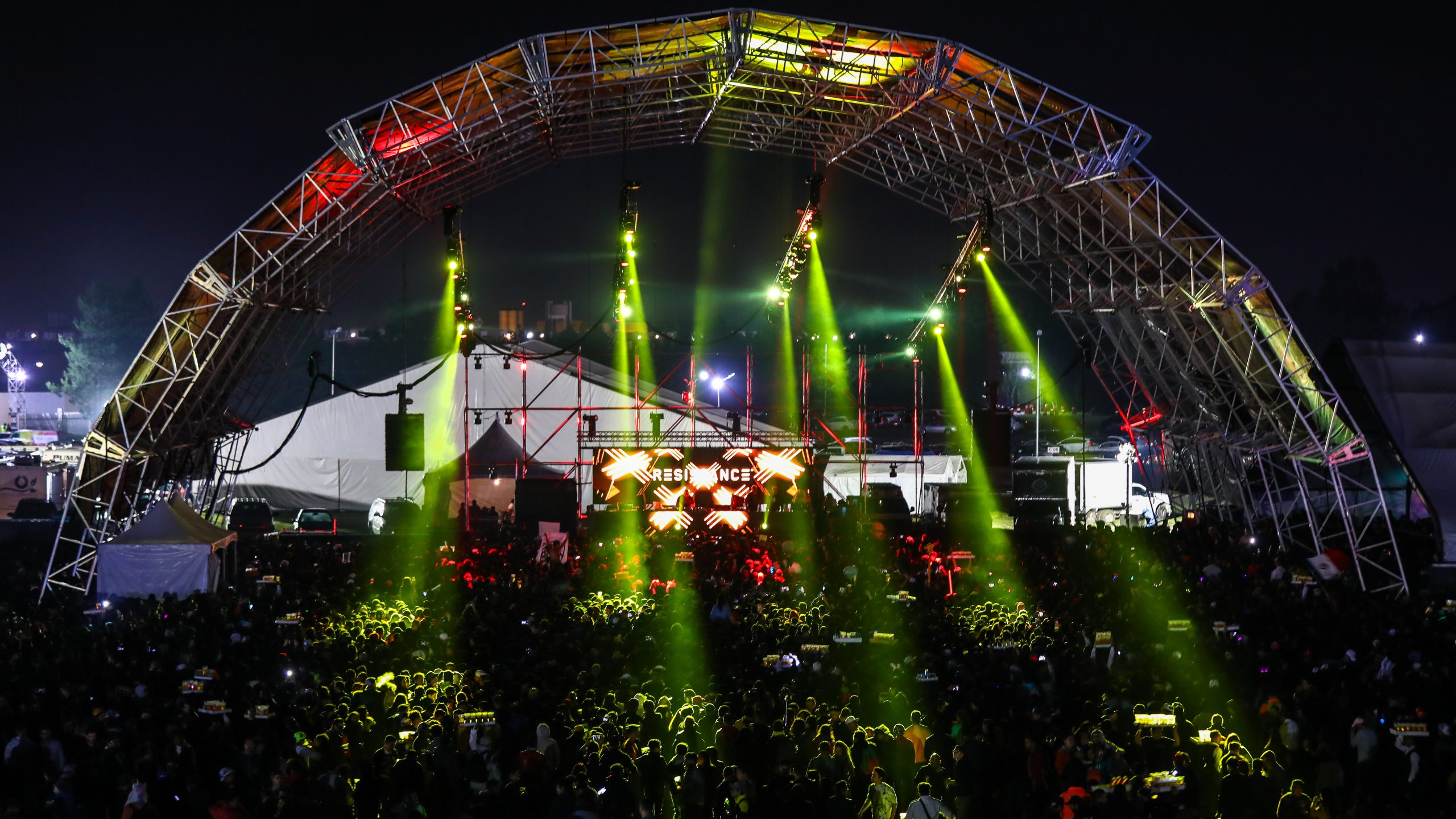 Escenario secundario de noche diseñado con un gran arco metálico iluminado por potentes reflectores de luz verde que caen en diagonal hacia la multitud congregada en el campo del festival.