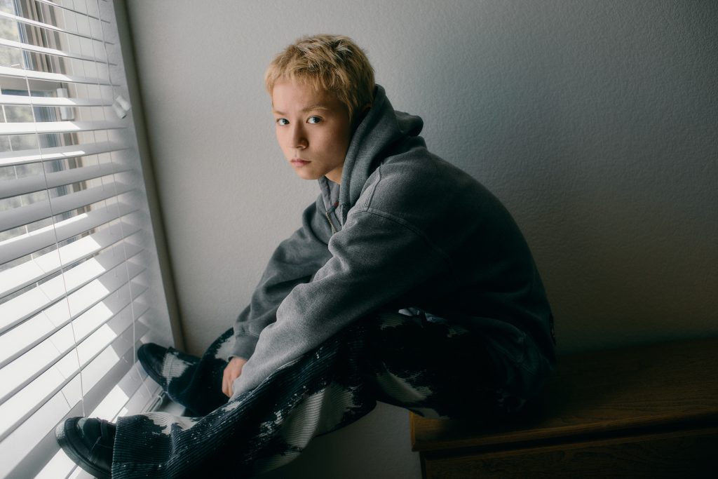 Rui Suzuki de KID PHENOMENON posa junto a una ventana con sudadera gris durante una sesión fotográfica.